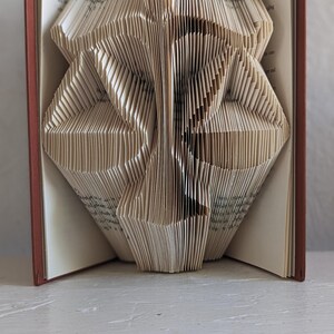 May include: A book with pages folded to create a three-dimensional sculpture of a scale of justice. The book has a dark red cover, and the interior pages are cream-colored with visible black printed text on the edges of the folded paper.