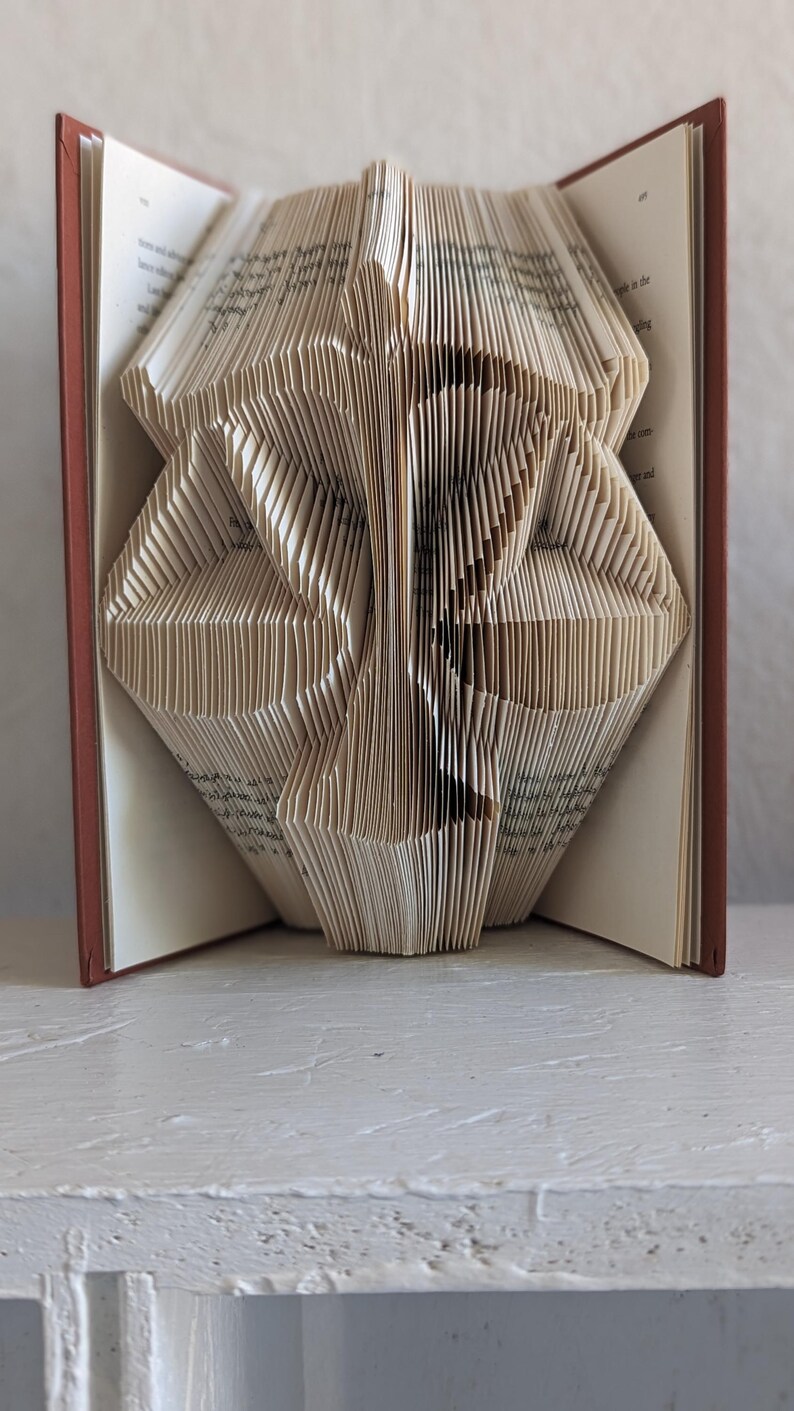 May include: A book with pages folded to create a three-dimensional sculpture of a scale of justice. The book has a dark red cover, and the interior pages are cream-colored with visible black printed text on the edges of the folded paper.