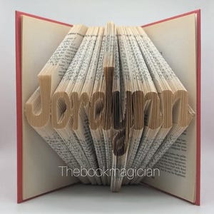May include: A book with the name "Jordyn" folded into the pages. The book is open and the pages are folded to create the name. The book has a red cover.