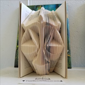 May include: A book sculpture featuring folded pages that form the shape of a scale of justice. The open book has cream-colored pages with black printed text. A small white card in front displays the text THEBOOKMAGICIAN with a book icon.