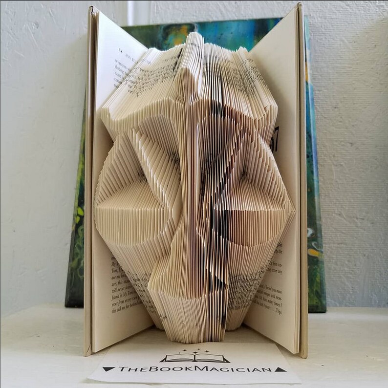 May include: A book sculpture featuring folded pages that form the shape of a scale of justice. The open book has cream-colored pages with black printed text. A small white card in front displays the text THEBOOKMAGICIAN with a book icon.