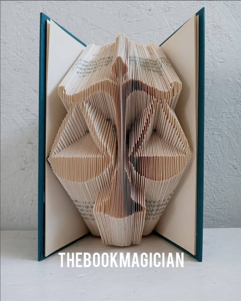 May include: A folded book sculpture featuring the shape of a scale of justice, with cream-colored pages and teal covers. The text THEBOOKMAGICIAN is printed in white at the bottom. The pages are precisely folded to create a three-dimensional silhouette.