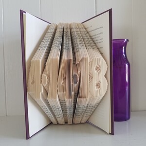 May include: A book with the word "HOME" folded into the pages. The book is open and the pages are folded to create the letters. The book is on a white surface with a purple vase to the right.