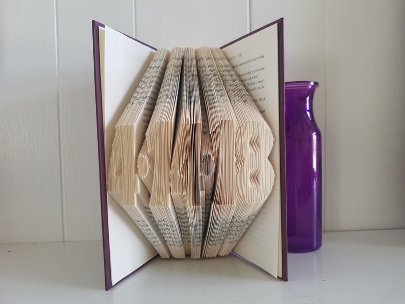 May include: A book with the word "HOME" folded into the pages. The book is open and the pages are folded to create the letters. The book is on a white surface with a purple vase to the right.