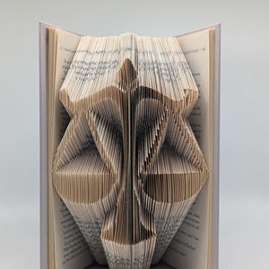 May include: A book with pages folded to create a three-dimensional sculpture of a set of scales of justice. The cream-colored paper pages are precisely creased to form the central pillar and two balanced pans of the scale symbol.