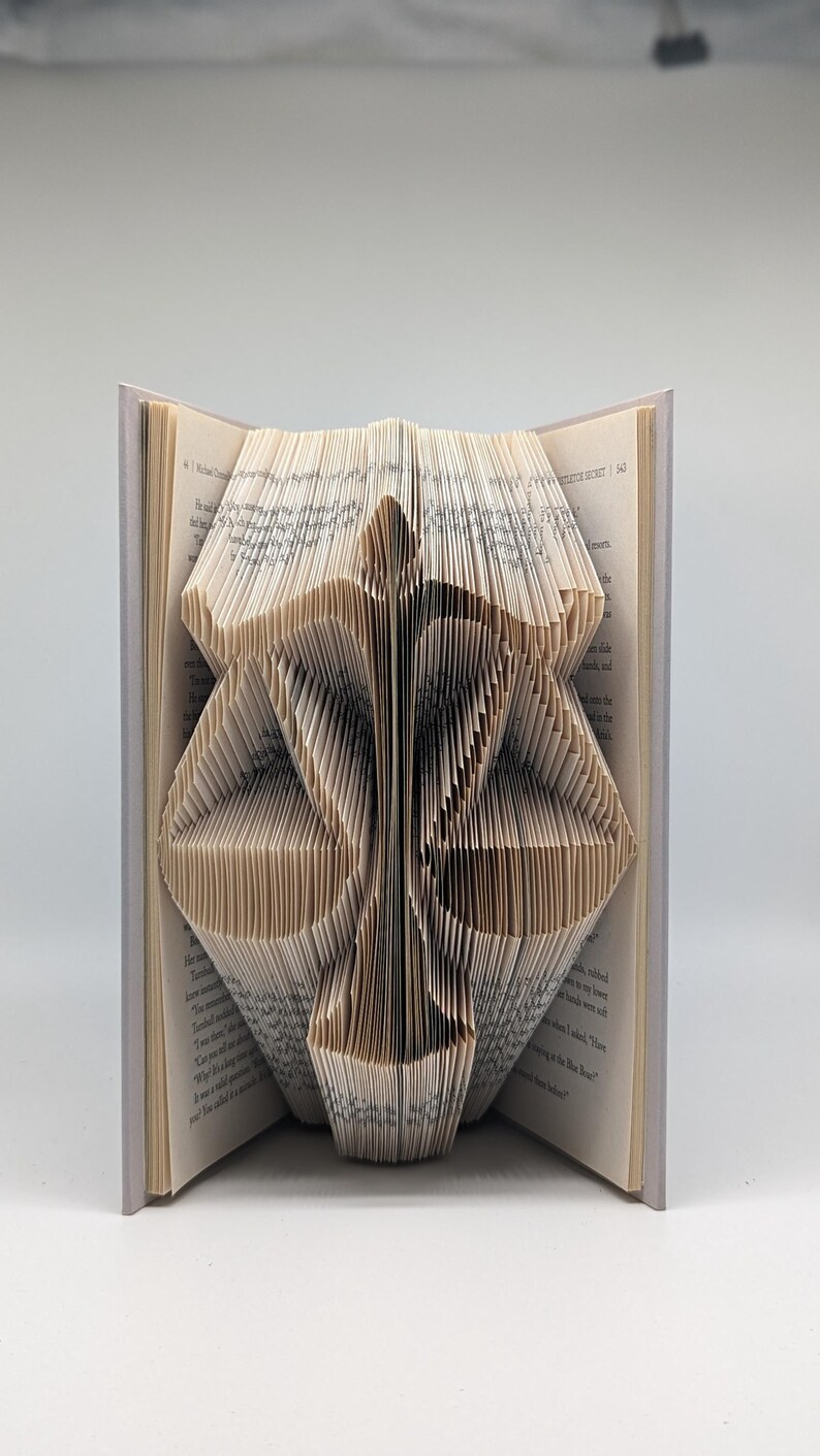 May include: A book with pages folded to create a three-dimensional sculpture of a set of scales of justice. The cream-colored paper pages are precisely creased to form the central pillar and two balanced pans of the scale symbol.