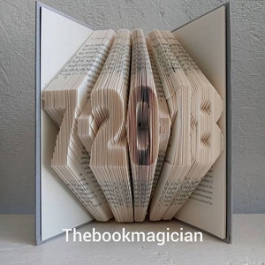 May include: A book art sculpture shaped like the numbers 12-20-12. The book is folded and fanned out to create the shape. The book is open and the pages are visible. The book is on a white surface. The book is titled "Thebookmagician".