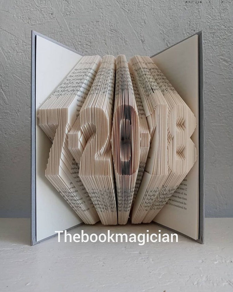 May include: A book art sculpture shaped like the numbers 12-20-12. The book is folded and fanned out to create the shape. The book is open and the pages are visible. The book is on a white surface. The book is titled "Thebookmagician".