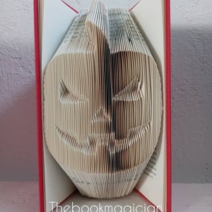 May include: A book folded into the shape of a jack-o'-lantern. The book has a red cover and the pages are folded to create a 3D effect. The book is standing upright on a white surface. The text "Thebookmagician" is printed on the bottom of the book.