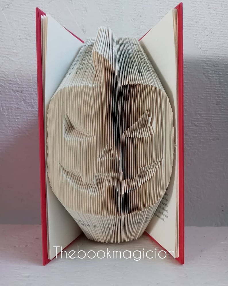 May include: A book folded into the shape of a jack-o'-lantern. The book has a red cover and the pages are folded to create a 3D effect. The book is standing upright on a white surface. The text "Thebookmagician" is printed on the bottom of the book.