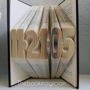 May include: A book with the year 1215 folded into the pages. The book is closed and standing upright on a white surface. The book is bound in black with the text "Thebookmagician" printed on the front cover.