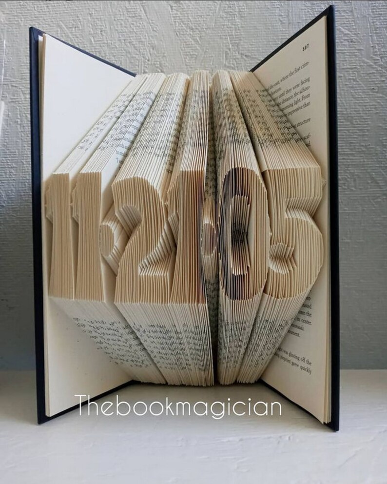 May include: A book with the year 1215 folded into the pages. The book is closed and standing upright on a white surface. The book is bound in black with the text "Thebookmagician" printed on the front cover.