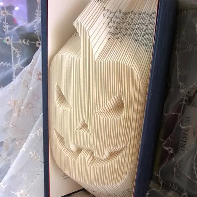 May include: A book folded into the shape of a jack-o'-lantern. The book is bound in a dark blue cover and the pages are cream colored.