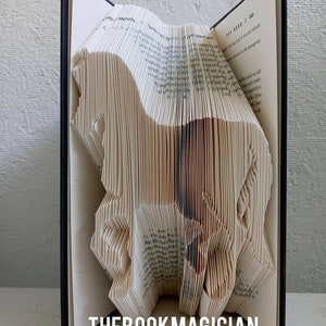 May include: A book art sculpture of a horse made by folding the pages of a book. The book is open and the pages are folded into a three-dimensional shape. The sculpture is standing on a white surface. The text "THEBOOKMAGICIAN" is printed on the bottom of the image.