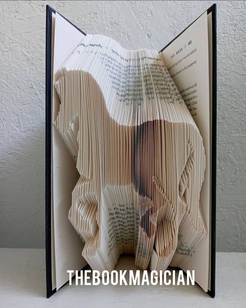 May include: A book art sculpture of a horse made by folding the pages of a book. The book is open and the pages are folded into a three-dimensional shape. The sculpture is standing on a white surface. The text "THEBOOKMAGICIAN" is printed on the bottom of the image.