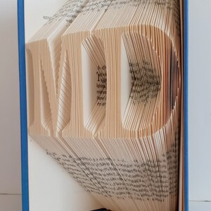 May include: A book with pages folded into the shape of the word "MID" in a blue book cover. The book is standing upright on a white surface.