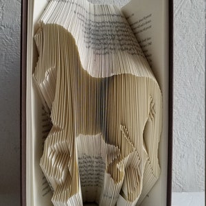 May include: A book art sculpture of a horse made by folding the pages of a book. The book is closed and has a brown cover. The sculpture is standing on a white surface. The text "THEBOOKMAGICIAN" is visible on a white card in the foreground.
