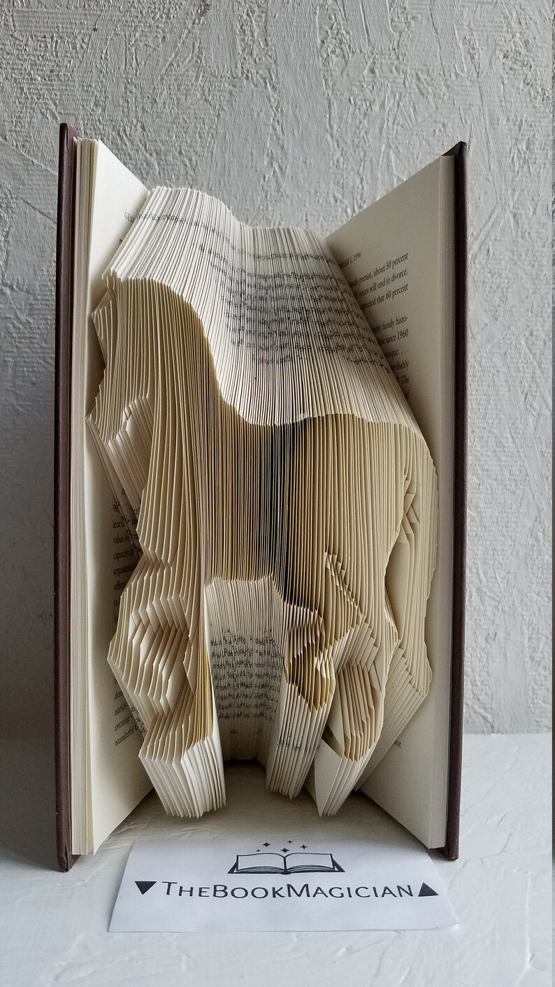 May include: A book art sculpture of a horse made by folding the pages of a book. The book is closed and has a brown cover. The sculpture is standing on a white surface. The text "THEBOOKMAGICIAN" is visible on a white card in the foreground.