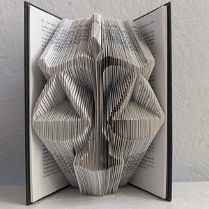 May include: A book with pages folded to create a three-dimensional sculpture of the scales of justice. The black hardback cover is visible on both sides, framing the intricate white paper folds that form the balanced scale shape.
