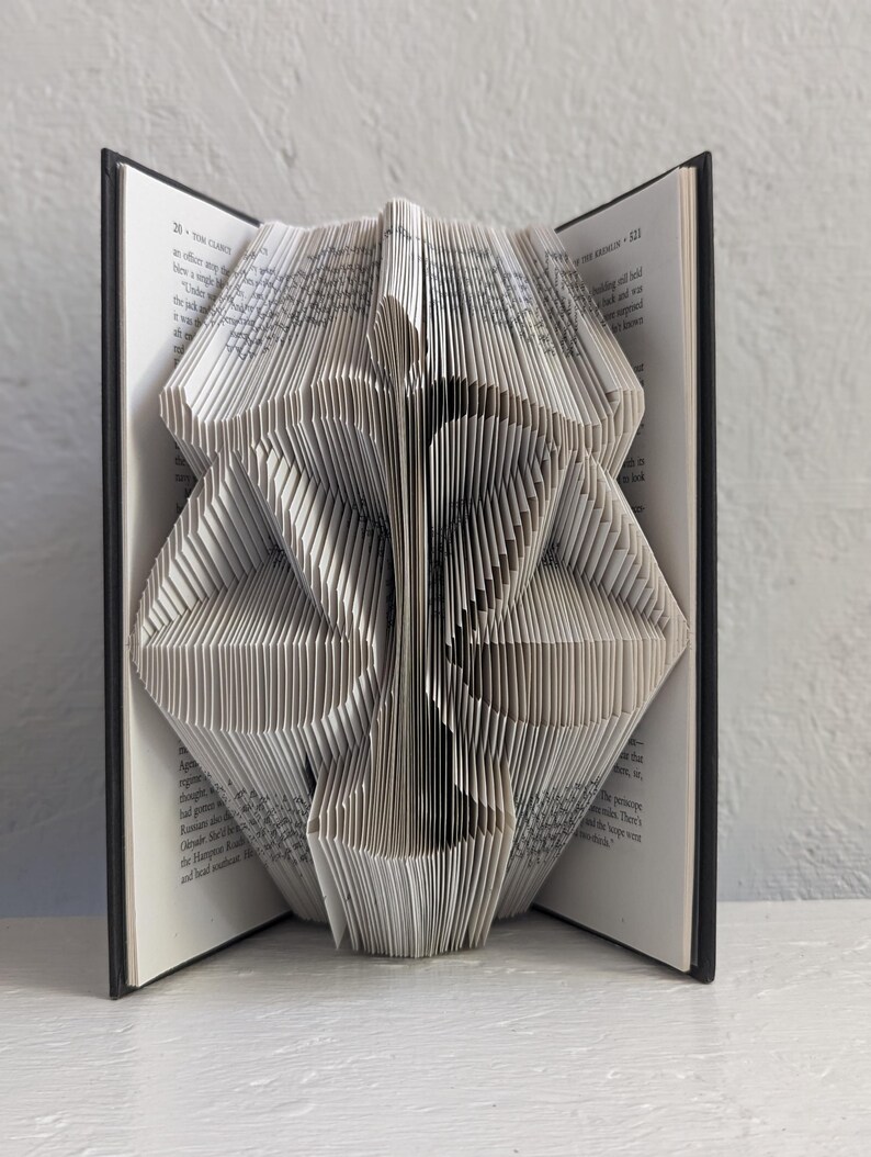 May include: A book with pages folded to create a three-dimensional sculpture of the scales of justice. The black hardback cover is visible on both sides, framing the intricate white paper folds that form the balanced scale shape.