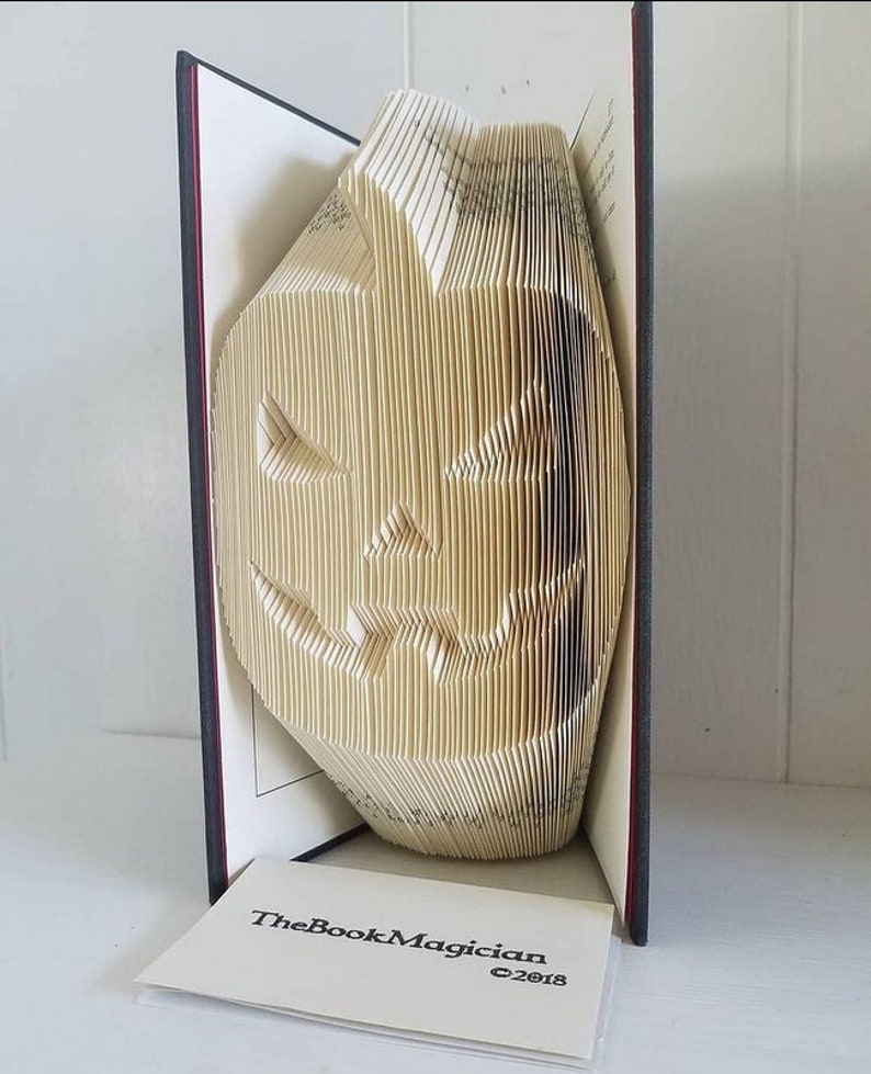 May include: A book folded into the shape of a jack-o'-lantern with a carved face. The book is standing upright on a white surface with a business card that reads "TheBookMagician &copy;2018".