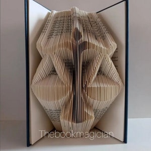 May include: A book sculpture featuring pages folded to create a symmetrical, abstract geometric shape. The hardback book has dark blue covers and white pages with visible text. The text "Thebookmagician" is printed in white at the bottom center.