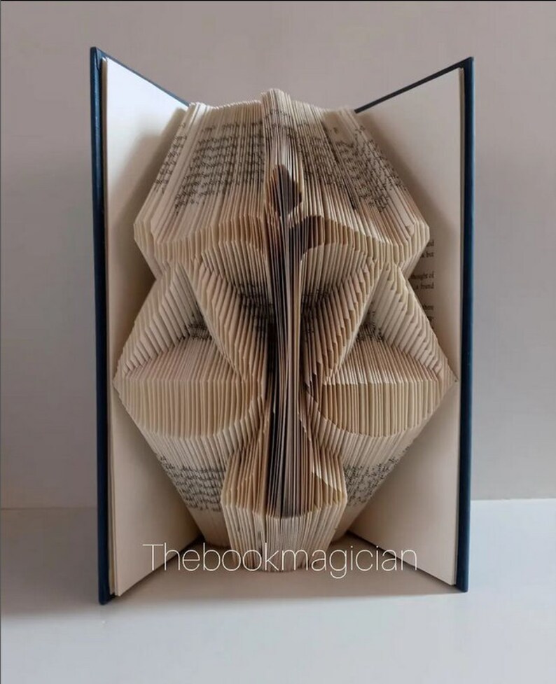 May include: A book sculpture featuring pages folded to create a symmetrical, abstract geometric shape. The hardback book has dark blue covers and white pages with visible text. The text "Thebookmagician" is printed in white at the bottom center.