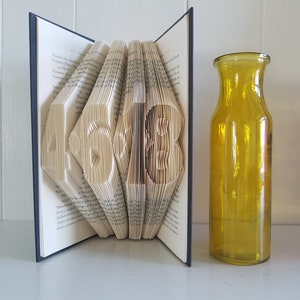 May include: A book with the numbers 45 and 13 folded into the pages. The book is open and standing upright. A yellow glass vase is to the right of the book.