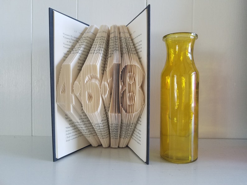 May include: A book with the numbers 45 and 13 folded into the pages. The book is open and standing upright. A yellow glass vase is to the right of the book.