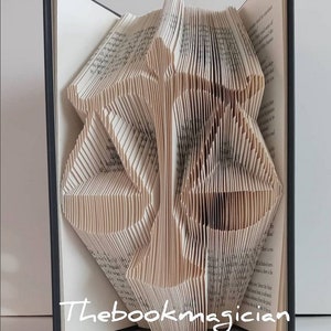 May include: A folded book sculpture featuring the shape of a scale of justice created by precisely folding the pages of a hardcover book. The white paper edges form the silhouette of the balance scale. The text Thebookmagician is visible.