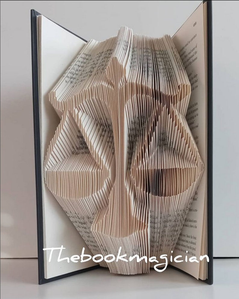 May include: A folded book sculpture featuring the shape of a scale of justice created by precisely folding the pages of a hardcover book. The white paper edges form the silhouette of the balance scale. The text Thebookmagician is visible.
