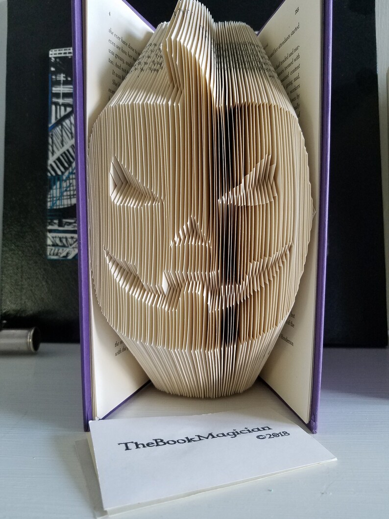 May include: A book folded into the shape of a jack-o'-lantern. The book is open and the pages are folded to create the shape of a face. The book is purple and has the text "TheBookMagician c2018" on the front.