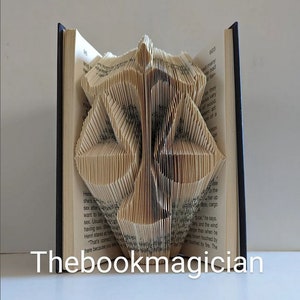 May include: A book sculpture featuring pages folded to create a symmetrical, geometric shape resembling a stylized mask or emblem. The open book has dark covers and visible printed text on the pages. The text Thebookmagician is displayed.
