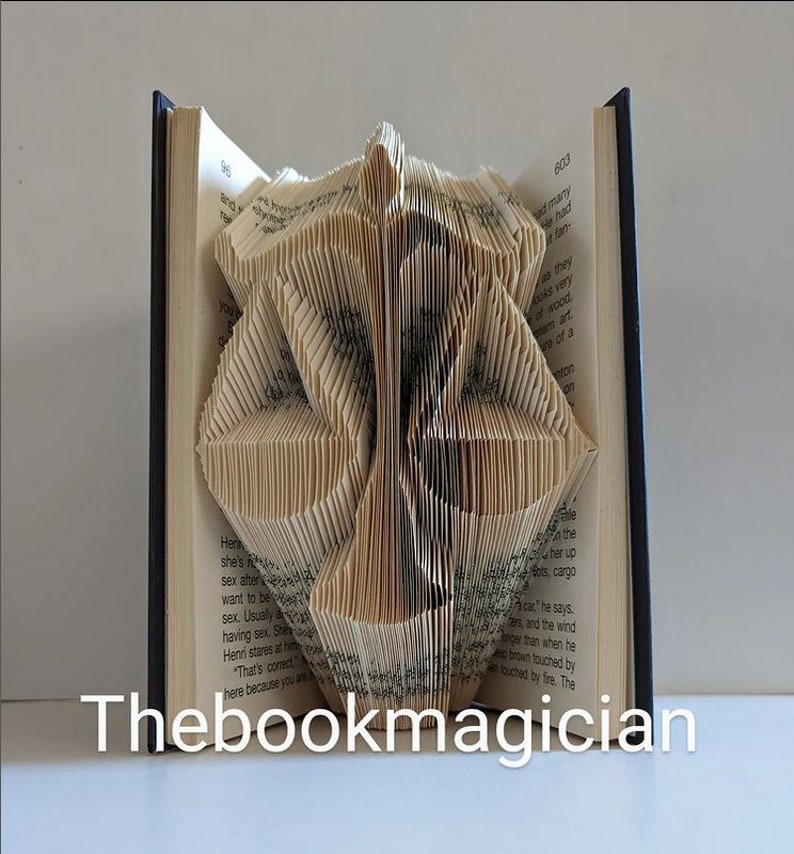 May include: A book sculpture featuring pages folded to create a symmetrical, geometric shape resembling a stylized mask or emblem. The open book has dark covers and visible printed text on the pages. The text Thebookmagician is displayed.