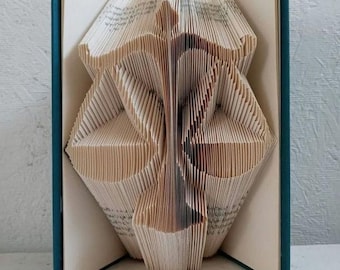 May include: A book sculpture with folded pages forming the shape of a scale of justice. The book has a dark teal cover and the text THEBOOKMAGICIAN is printed in white at the bottom. The paper pages are cream-colored and intricately folded.