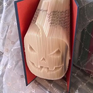 May include: A book art sculpture of a jack-o'-lantern made from the pages of a book. The book is bound in red and black fabric and is open to reveal the folded pages that form the jack-o'-lantern.