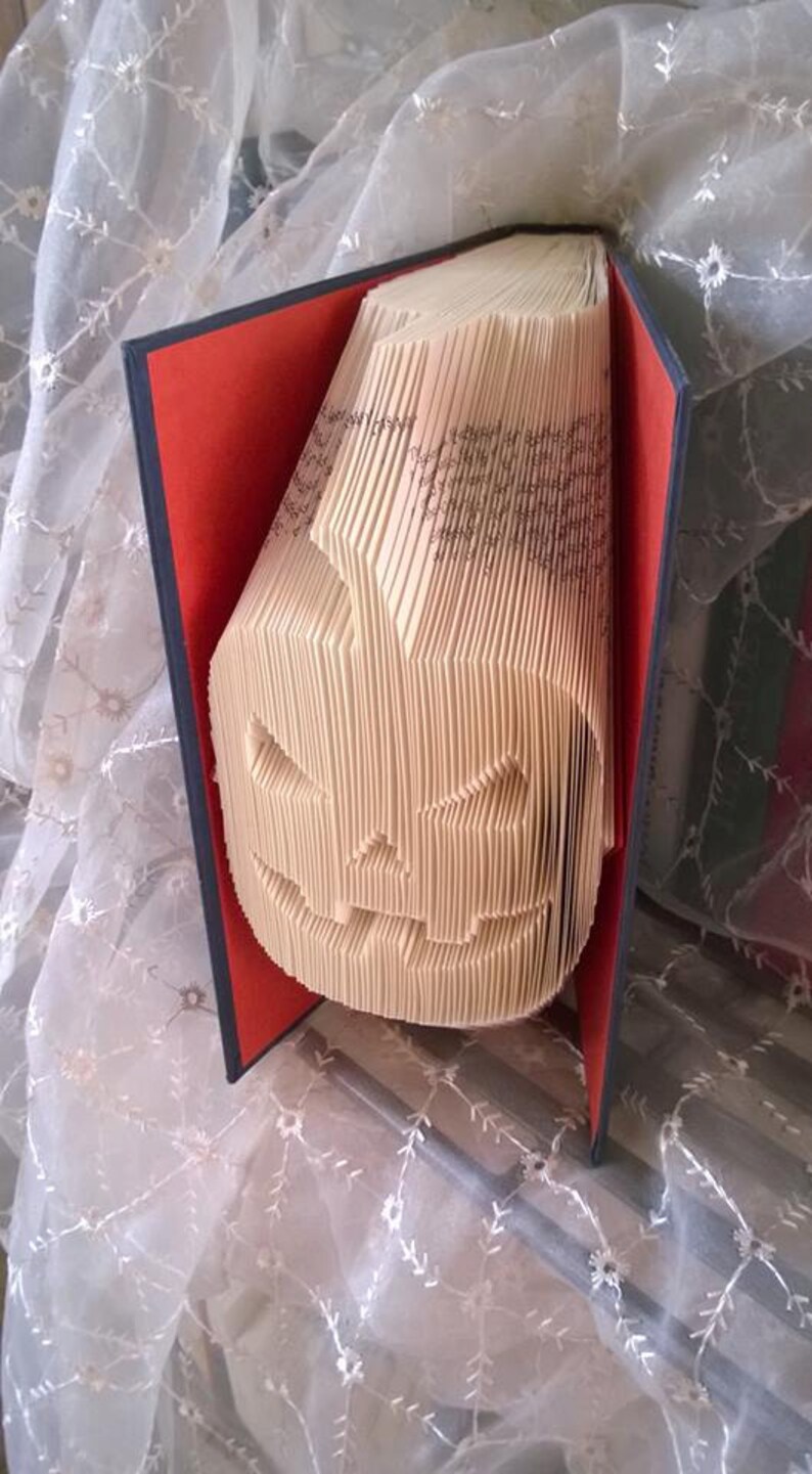May include: A book art sculpture of a jack-o'-lantern made from the pages of a book. The book is bound in red and black fabric and is open to reveal the folded pages that form the jack-o'-lantern.