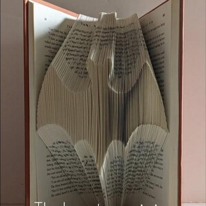 May include: A book folded into the shape of a Batman logo. The book is open and the pages are folded to create the shape. The book is brown with a white spine.