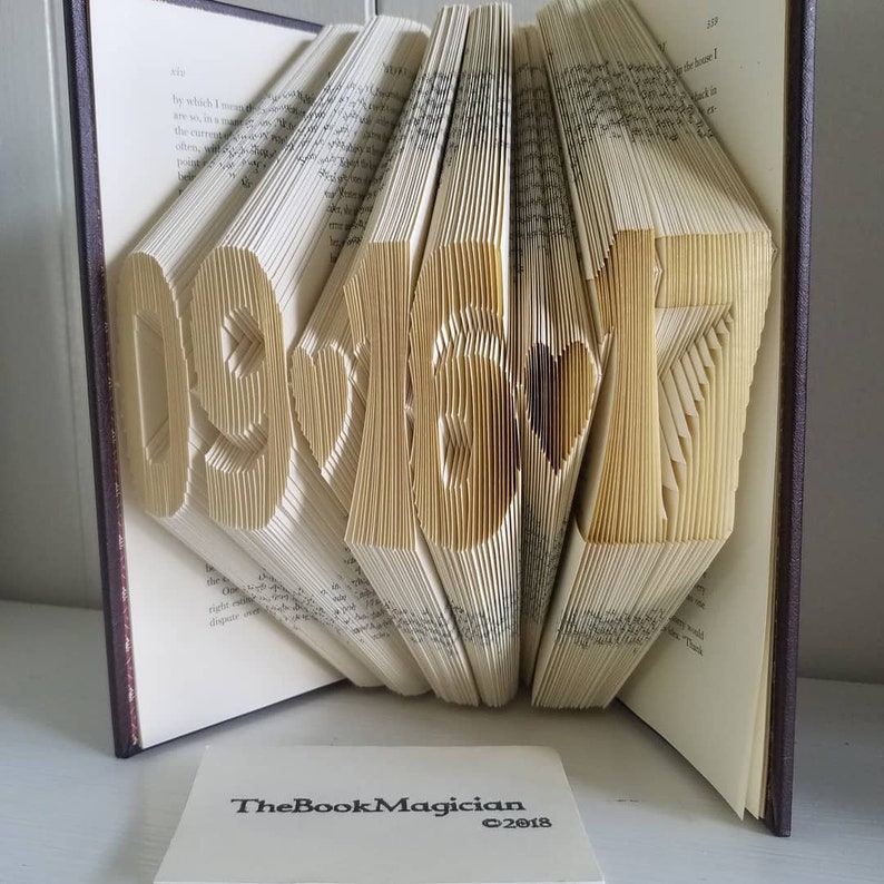 May include: A book with the date 091617 folded into the pages. The book is open and the pages are folded to create the date. The book is brown and has a gold spine. The book is sitting on a white surface. The book has a business card that says "TheBookMagician &copy;2018" on it.