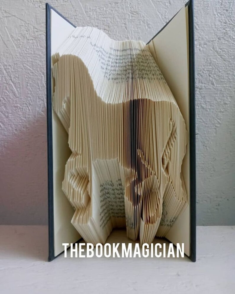 May include: A book art sculpture of a horse, folded from the pages of a book. The book is open and the pages are folded into the shape of a horse. The book is closed on the left and right sides, and the spine is visible. The book is on a white surface. The text "THEBOOKMAGICIAN" is visible at the bottom of the image.