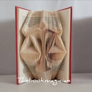 May include: A book with pages folded to create a three-dimensional sculpture of a scale of justice. The book has a red cover and the text Thebookmagician is printed in white at the bottom. The folded pages form the shape of a balanced scale.