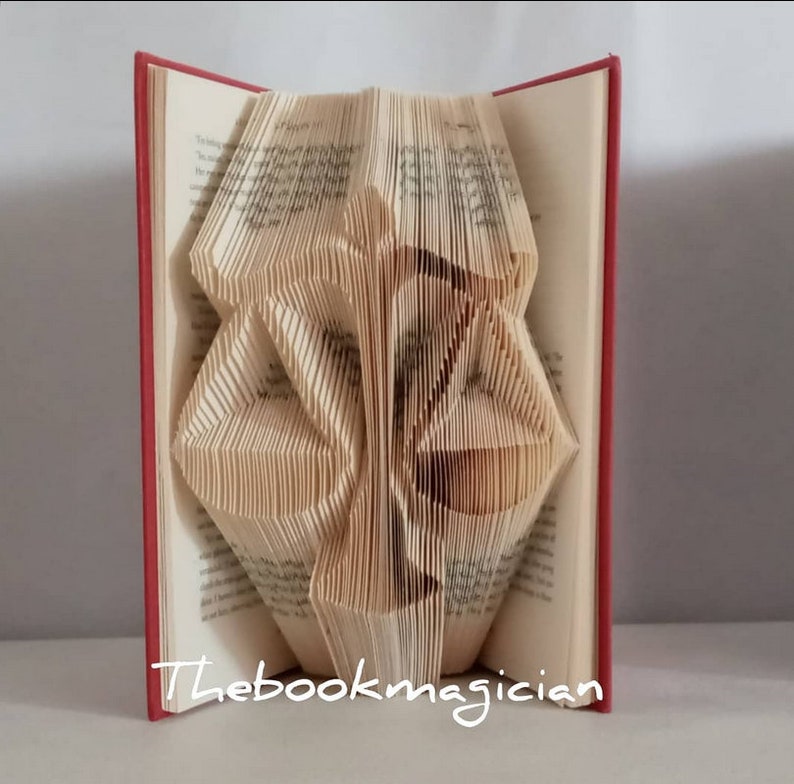 May include: A book with pages folded to create a three-dimensional sculpture of a scale of justice. The book has a red cover and the text Thebookmagician is printed in white at the bottom. The folded pages form the shape of a balanced scale.