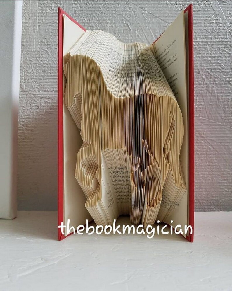 May include: A book art sculpture of a horse, folded from the pages of a book with red covers. The book is standing upright on a white surface. The text "thebookmagician" is printed on the bottom of the book.