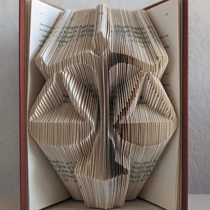 May include: A book with pages folded to create a three-dimensional sculpture of the scales of justice. The book has a dark red cover and white pages with visible black printed text. The folded paper forms a symmetrical shape resembling a balance scale.