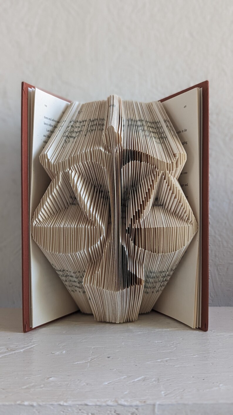 May include: A book with pages folded to create a three-dimensional sculpture of the scales of justice. The book has a dark red cover and white pages with visible black printed text. The folded paper forms a symmetrical shape resembling a balance scale.