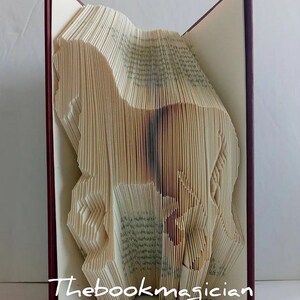May include: A book art sculpture of a horse, folded from the pages of a book. The book is bound in a burgundy cover and the sculpture is standing upright. The text "Thebookmagician" is visible on the bottom of the book.