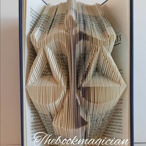 May include: A folded book sculpture featuring the silhouette of the scales of justice, created by precisely folding the pages of a hardcover book. The text Thebookmagician is printed in white script at the bottom of the pages.