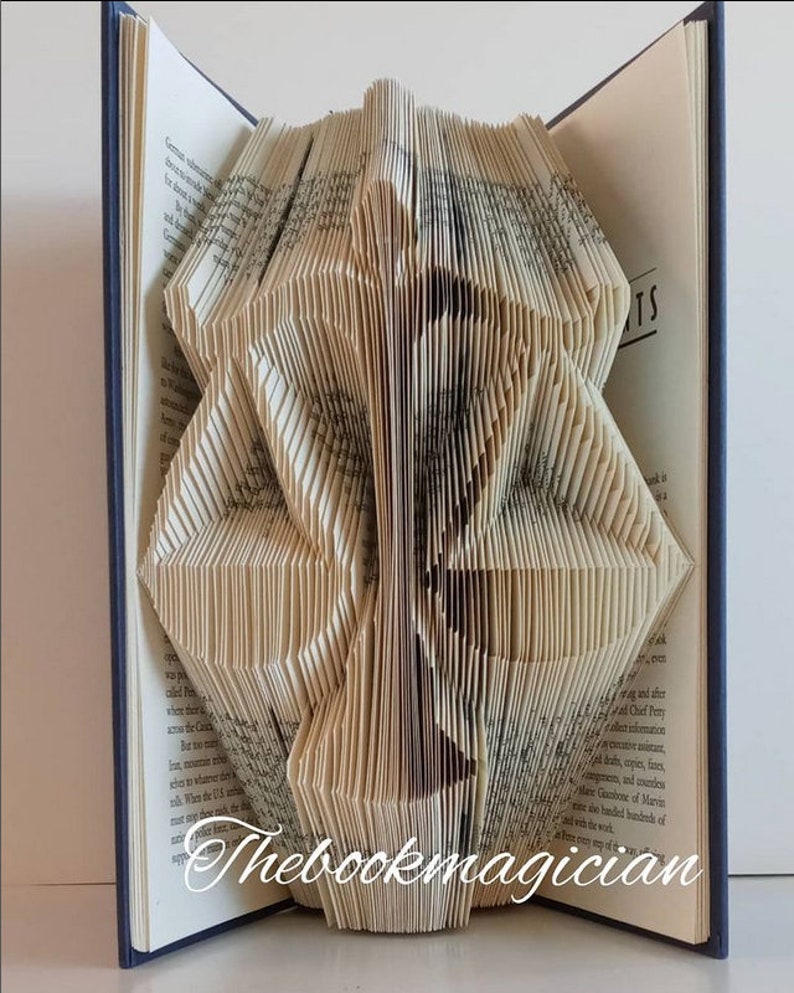 May include: A folded book sculpture featuring the silhouette of the scales of justice, created by precisely folding the pages of a hardcover book. The text Thebookmagician is printed in white script at the bottom of the pages.