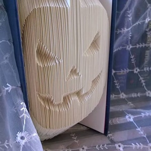 May include: A book folded into the shape of a jack-o'-lantern. The book is open and the pages are folded to create the shape of a face with a wide smile and two eyes. The book is sitting on a white lace tablecloth.