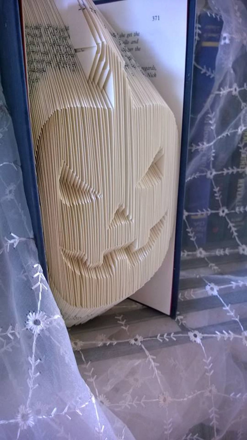 May include: A book folded into the shape of a jack-o'-lantern. The book is open and the pages are folded to create the shape of a face with a wide smile and two eyes. The book is sitting on a white lace tablecloth.